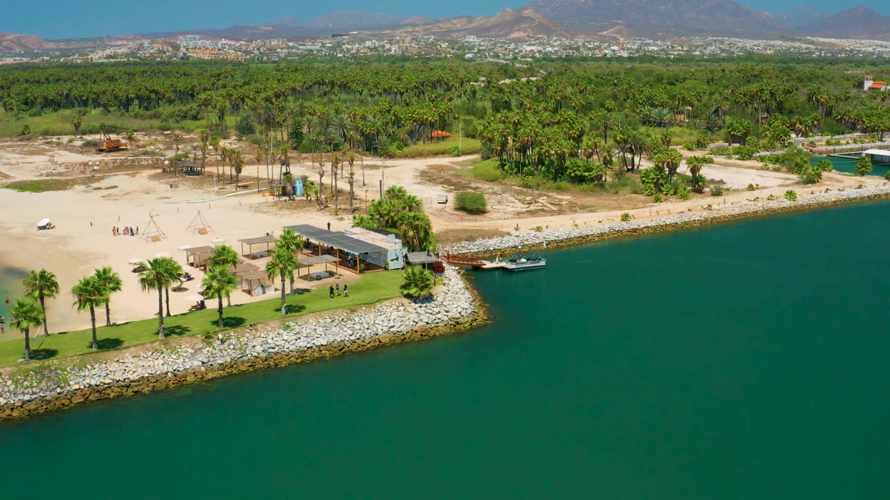Aerial frame in San Jose Del Cabo, Mexico, with the beach structure closer to center, palms along the shore, green water at right, sandy open land behind, and tiny beachgoers visible.