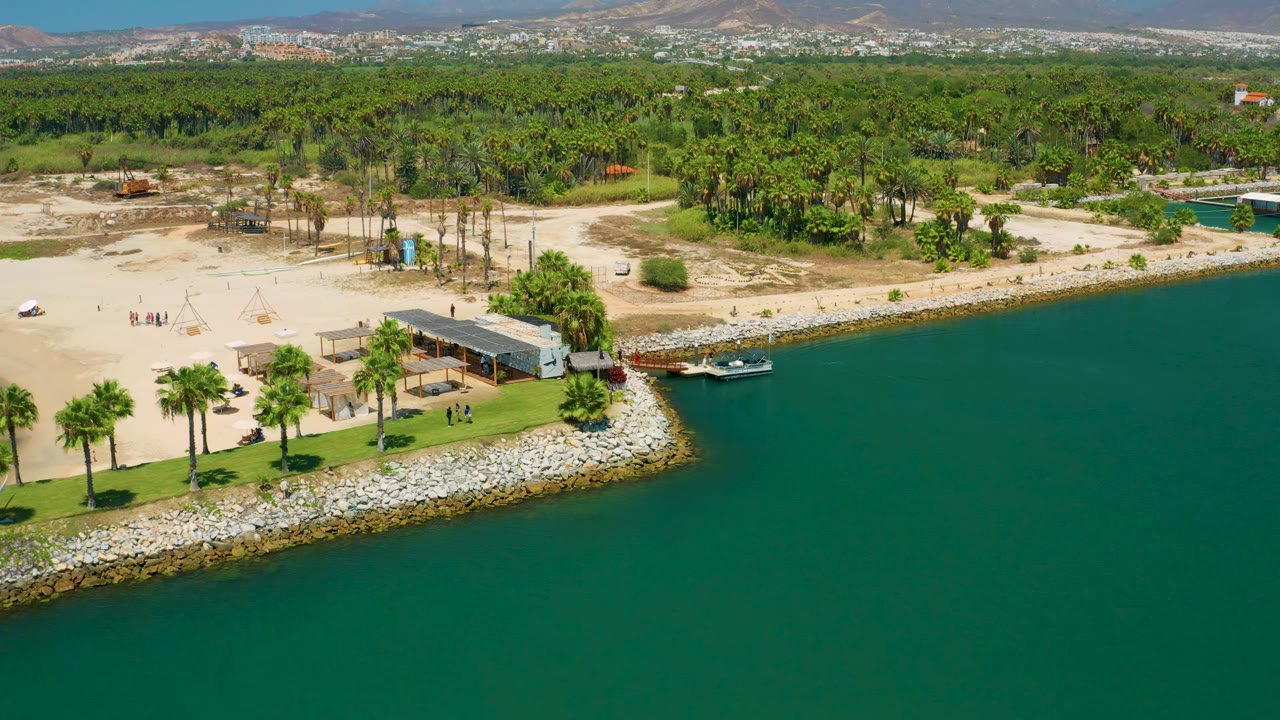 Aerial view in San Jose Del Cabo, Mexico, showing more open green water in the foreground, a beach facility and palms along the shore, sandy ground inland, distant hills, and tiny beachgoers visible.