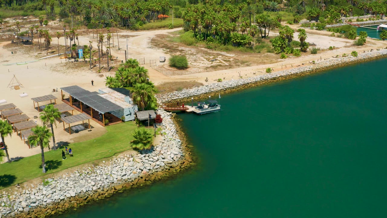 San Jose Del Cabo aerial frame with the small boat and pier centered near the seawall, green water on the right, sandy paths behind the shore, palms, pergola roofs, and tiny distant people on land.