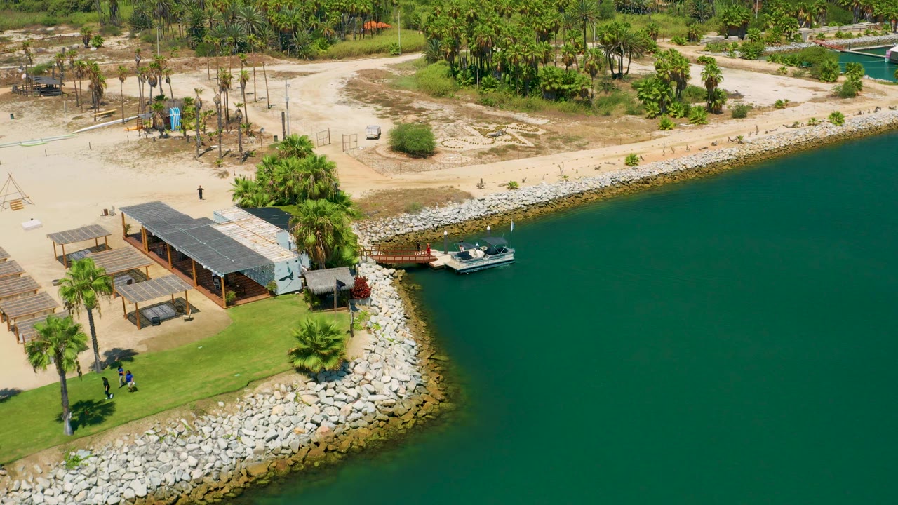 Aerial view in San Jose Del Cabo with the moored boat slightly farther right of the pier, a curved rock shoreline, palm clusters, shaded beach structures, sandy ground, and tiny distant people visible.