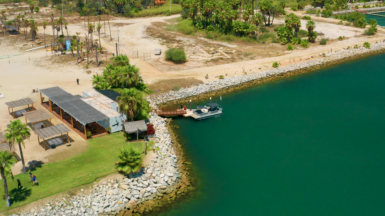 High-angle coastline with green water, a boat, pier, rocks, palms, and low buildings.