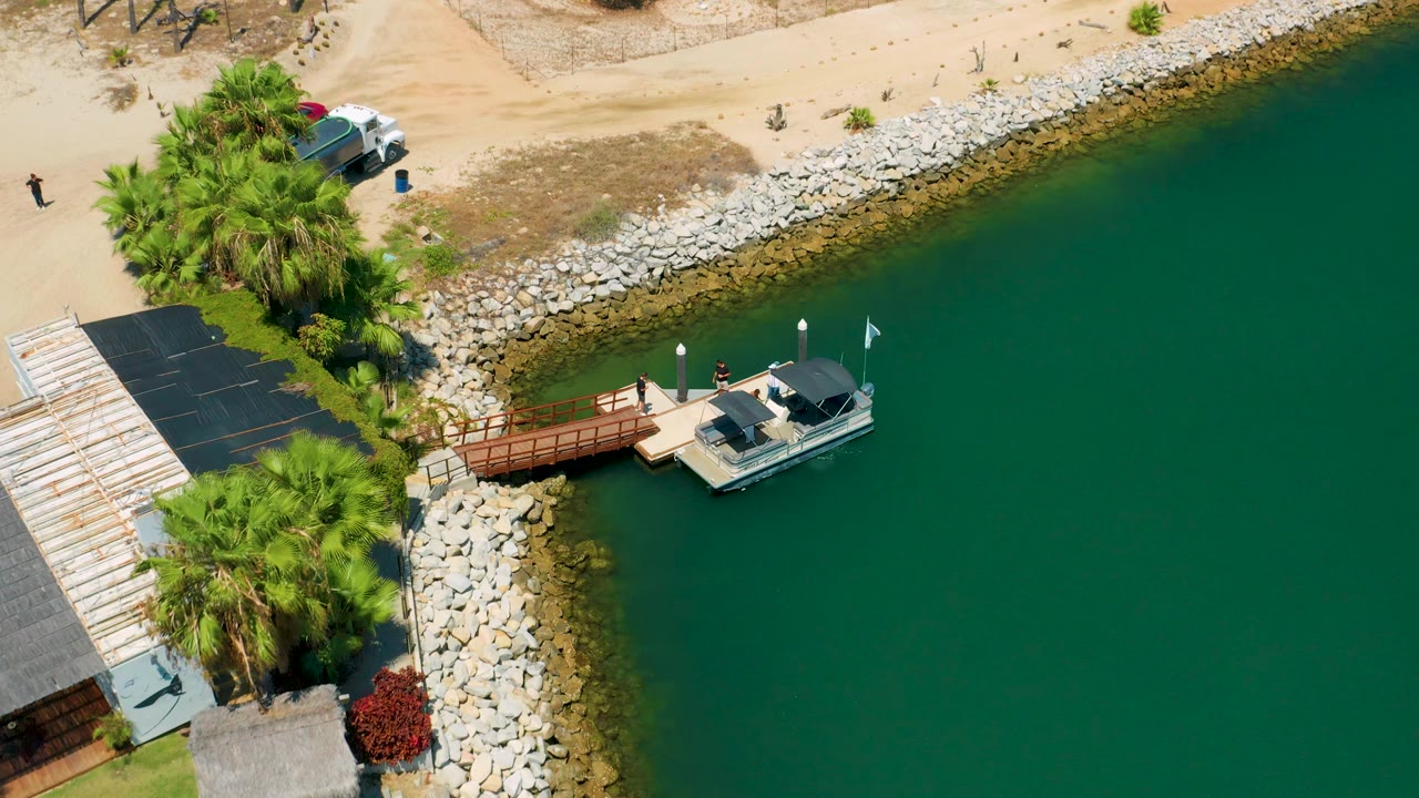 Overhead San Jose Del Cabo frame showing more open green water around the docked boat, with a sandy road, parked vehicles, palms, and visible people reduced to small figures.