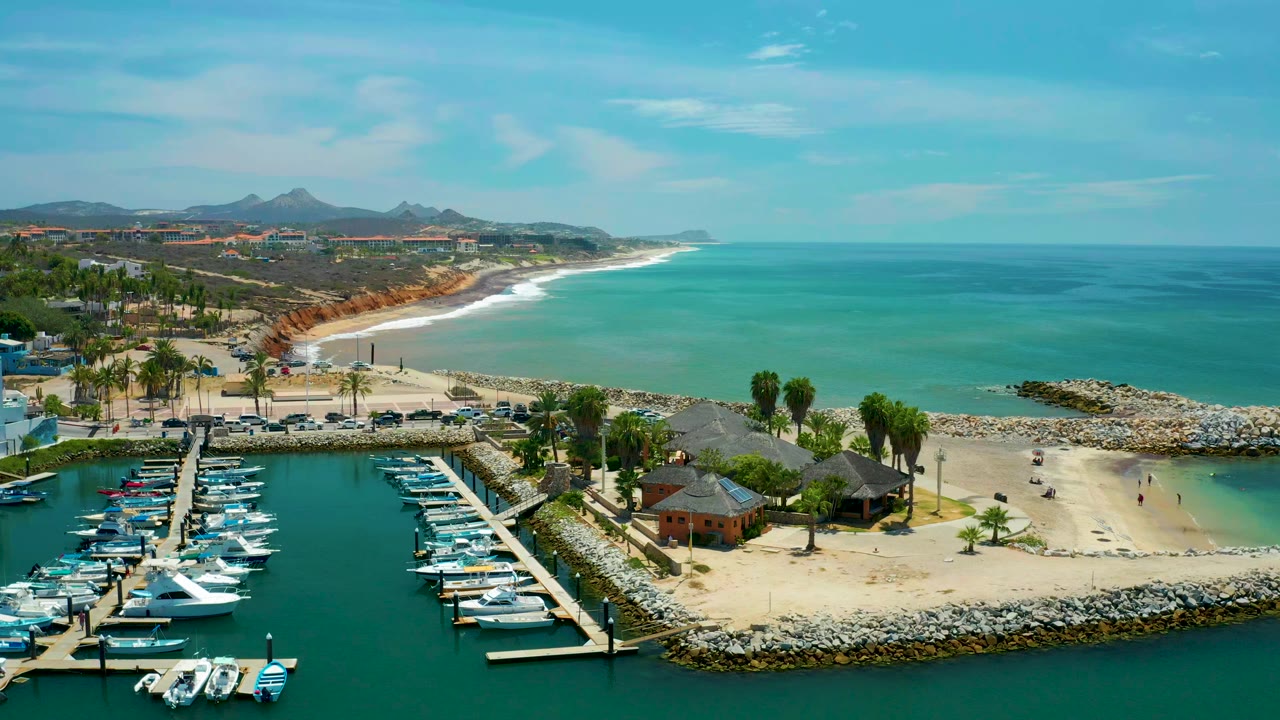 Aerial view over San Jose Del Cabo showing marina docks and boats in the foreground, a sandy beach and rock breakwater in the middle, turquoise water beyond, and no visible people.