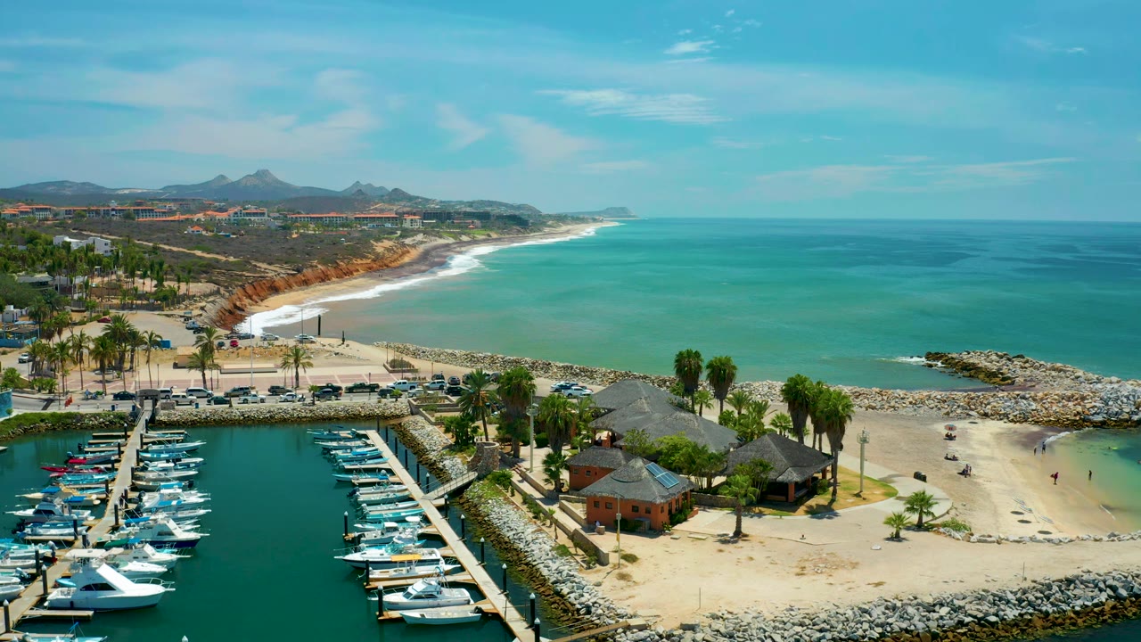 San Jose Del Cabo aerial frame with white boats lined along marina docks, palm trees and hut-like resting houses near the sand, rolling surf, open ocean, and no visible people.
