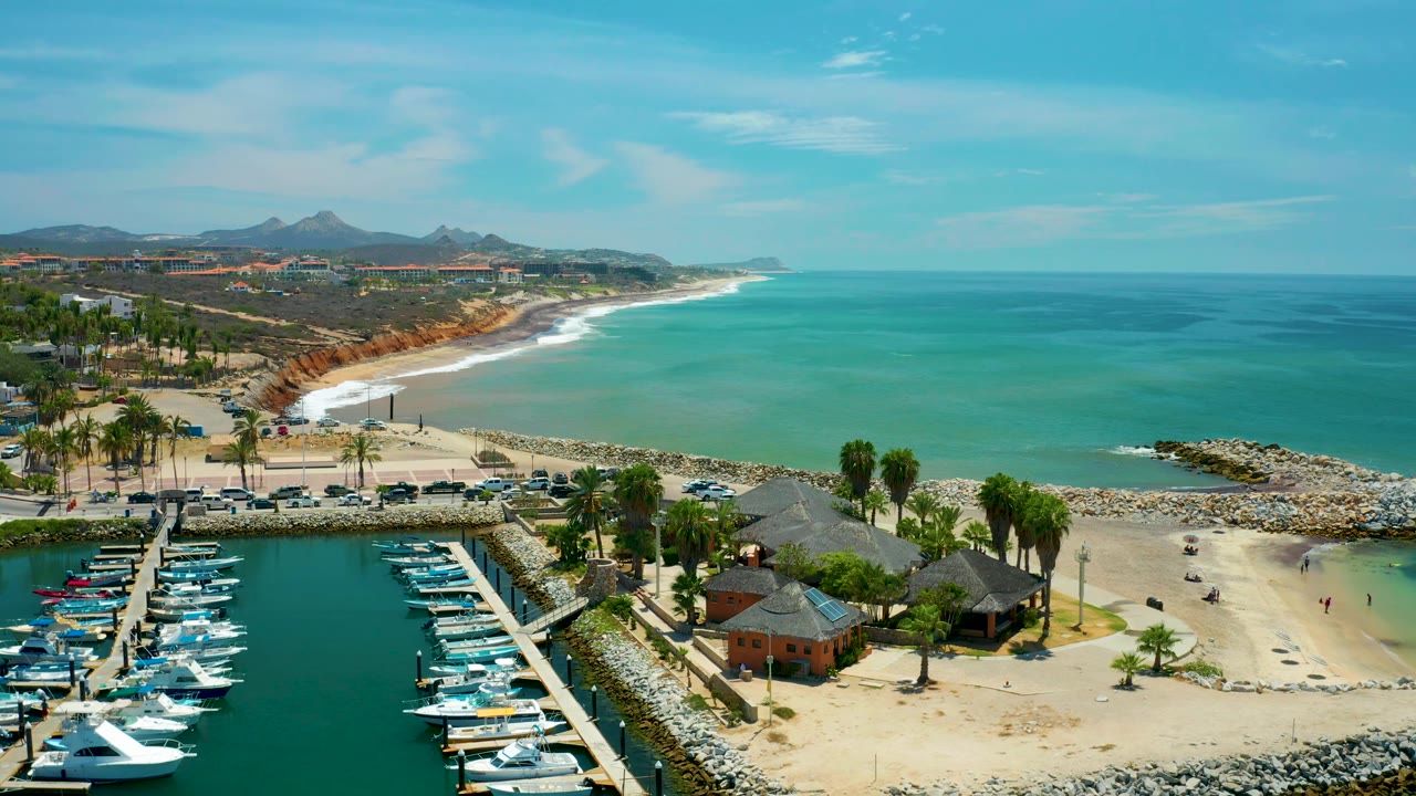 High aerial view in San Jose Del Cabo, Mexico with boats in the lower left marina, palm-lined sand and hut roofs near center, a rocky breakwater, blue-green ocean, and no visible people.