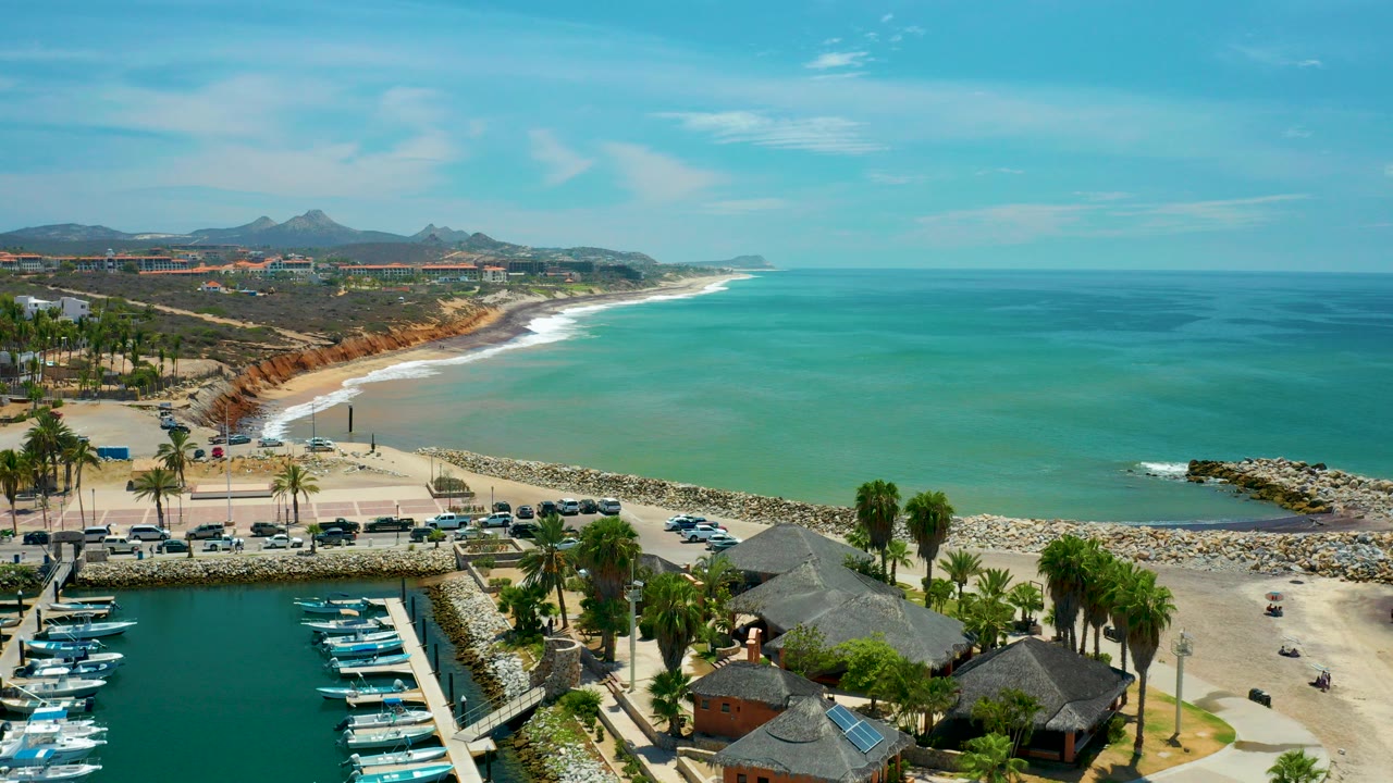 Wide aerial frame over San Jose Del Cabo, Mexico showing marina slips with small boats, palm trees, parked cars, a curved beach, turquoise water, distant hills, and no visible people.