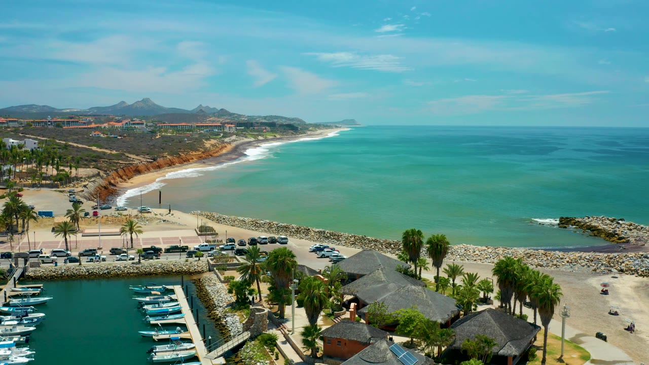 Wide daytime aerial over San Jose Del Cabo with thatched resort roofs and palms at lower right, parked cars near the beach, a rock breakwater, open turquoise sea, distant coastline, and no visible people.