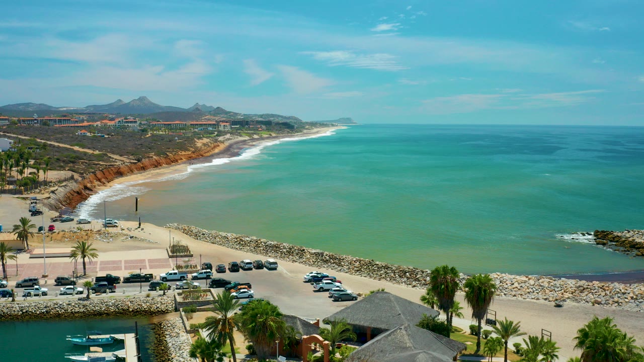 Wide aerial frame of San Jose Del Cabo coastline with a sandy beach, green-blue sea, rocky breakwater, road, parked cars, palms, distant hills, and no visible people.
