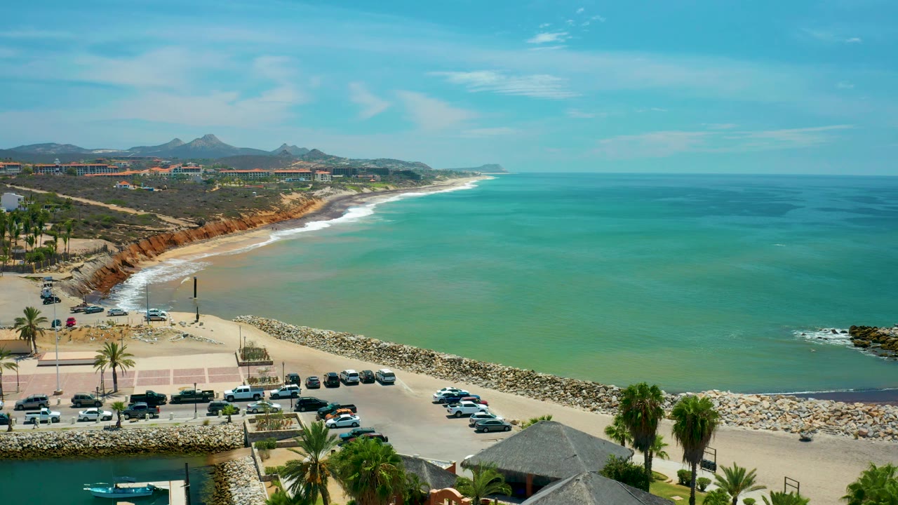 Elevated San Jose Del Cabo coastal view with green ocean filling the right side, sandy beach and surf to the left, breakwater below, roadway and cars in the foreground, and no visible people.