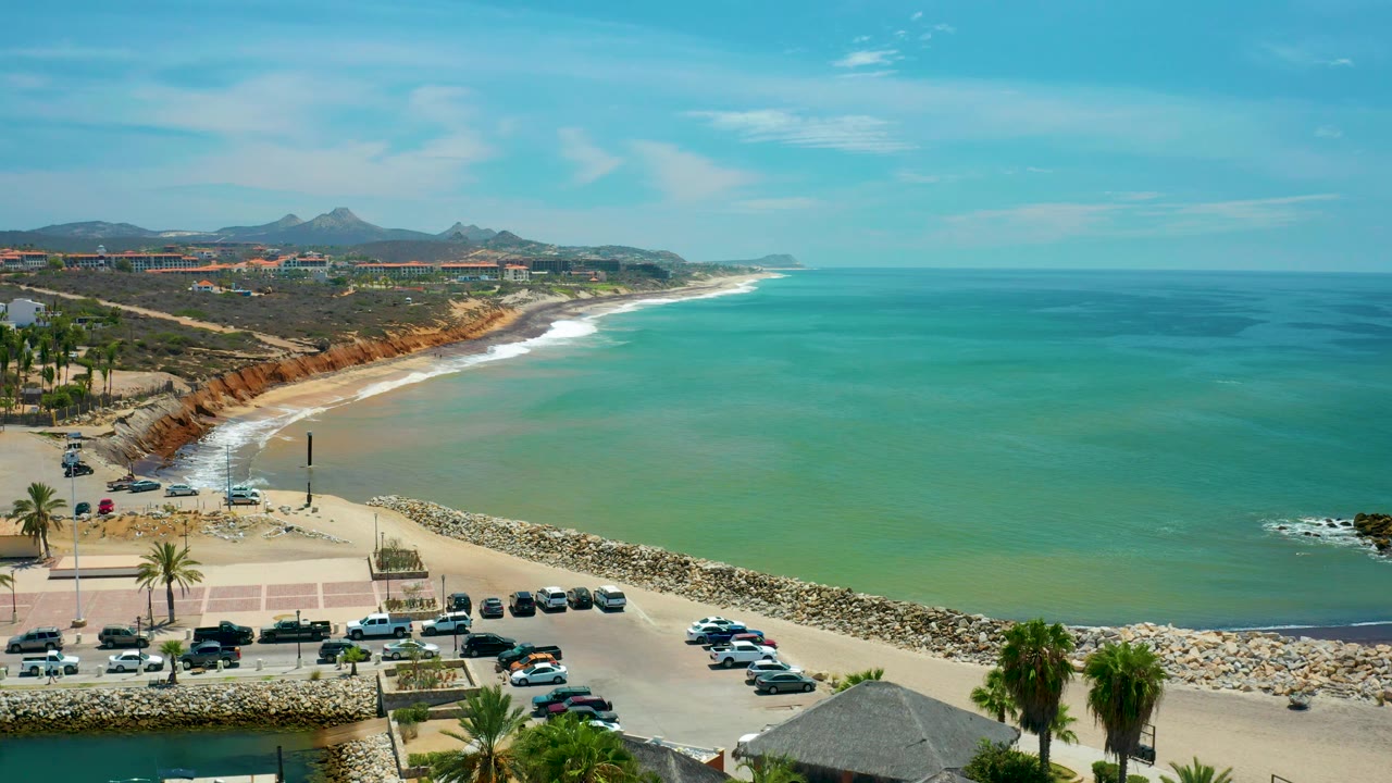 San Jose Del Cabo beach cove with green-blue water, a white wave band along the shore, rock breakwater across the lower frame, parked cars and palms below, and no visible people.