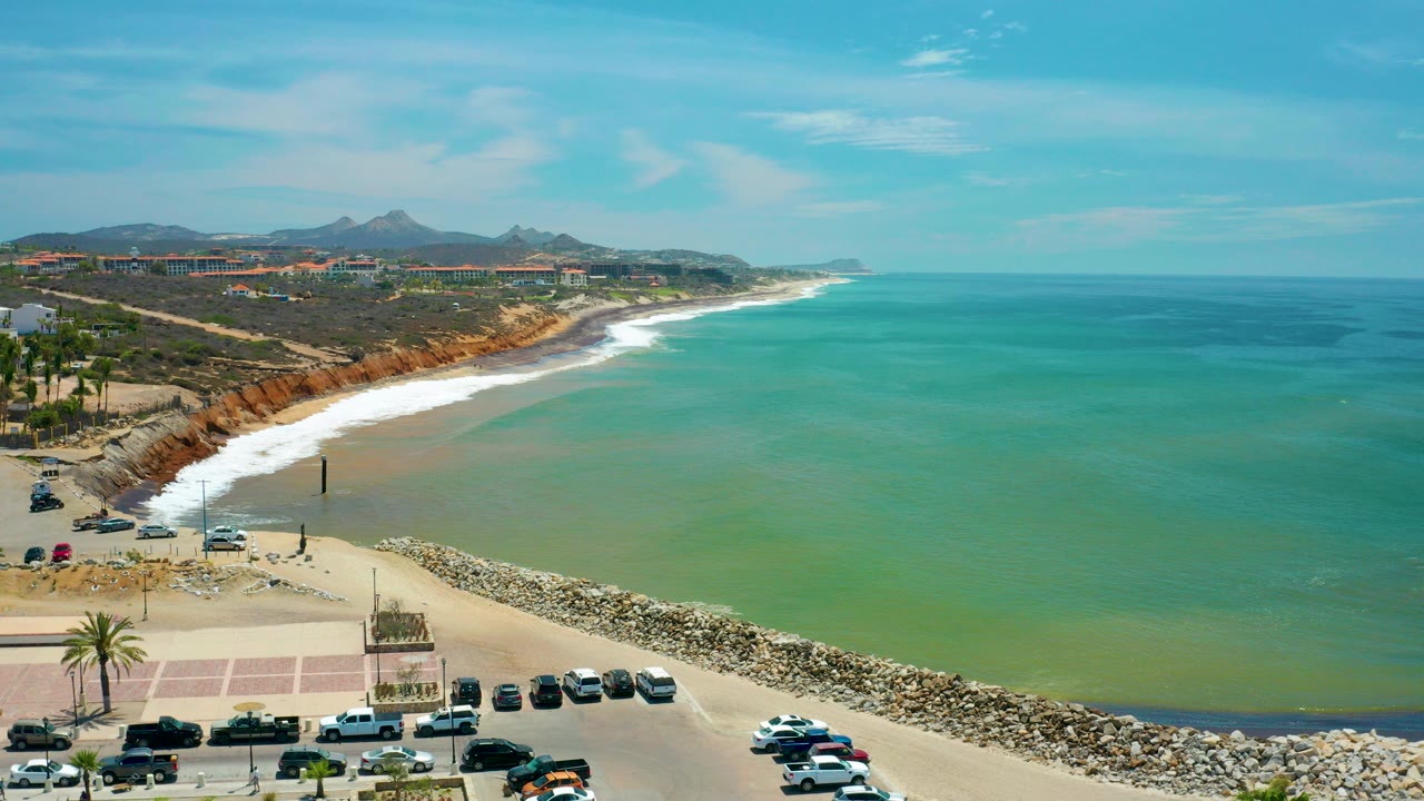 Wide aerial frame of San Jose Del Cabo coastline with parked cars at the lower edge, a rocky breakwater, pale sand, green-blue water, distant hills, and no visible people.