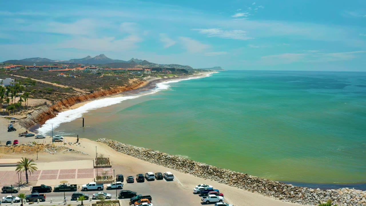 Aerial view over San Jose Del Cabo beach with a row of parked cars in the foreground, waves along the sand, reddish bluffs behind the beach, distant mountains, and no visible people.
