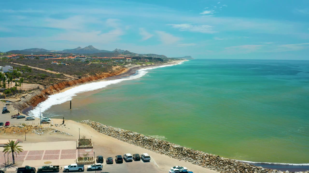 Aerial coastline with rock jetty, parked vehicles, sandy beach, and calm blue water.