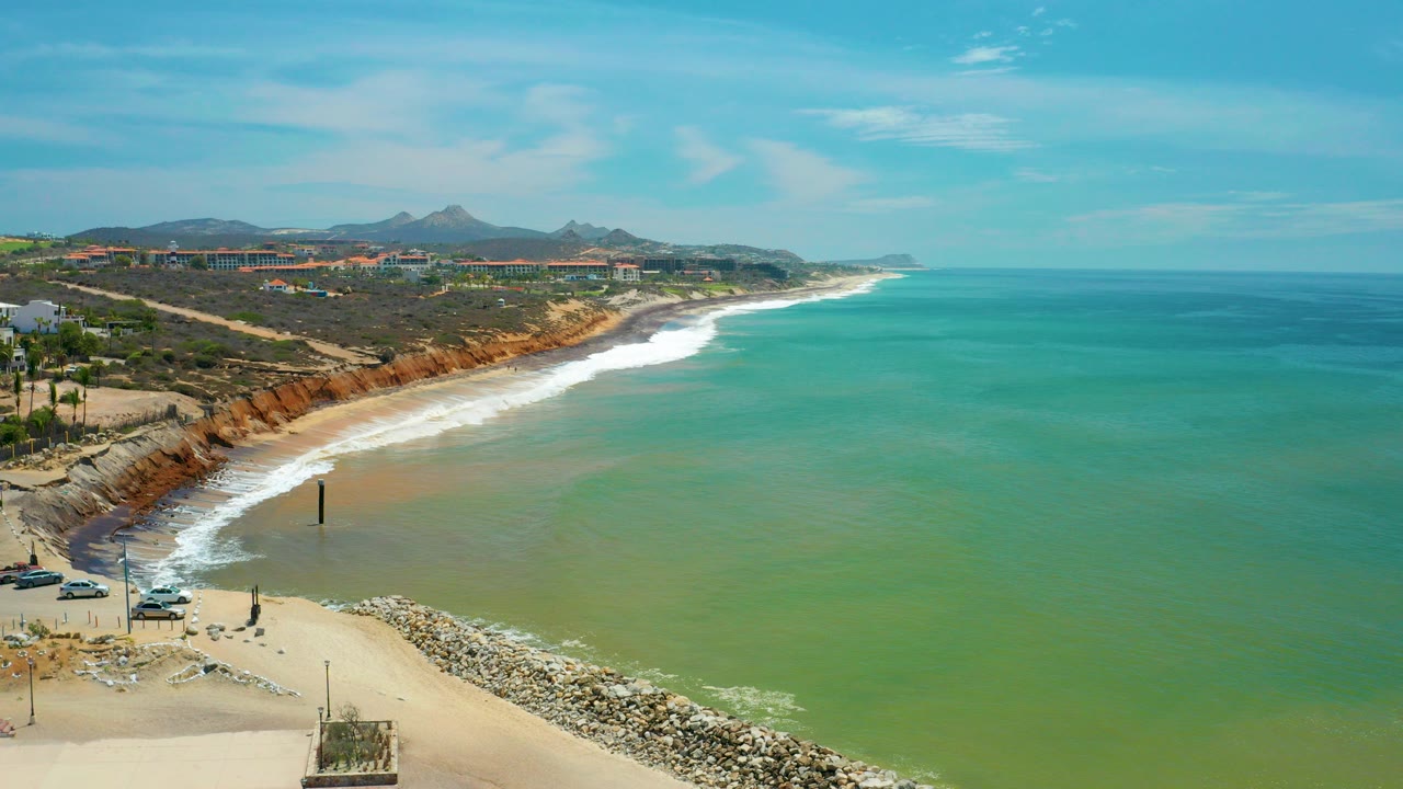Wide aerial frame over San Jose del Cabo with green-blue water, a curving surf line, sandy beach, rock jetty, rust bluffs, distant buildings, mountains, and no visible people.