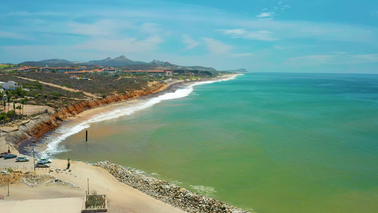 Aerial coastal view in San Jose del Cabo showing green water spread across the bay, a white wave band at the beach, the rock jetty below, bluffs, distant buildings, mountains, and no visible people.