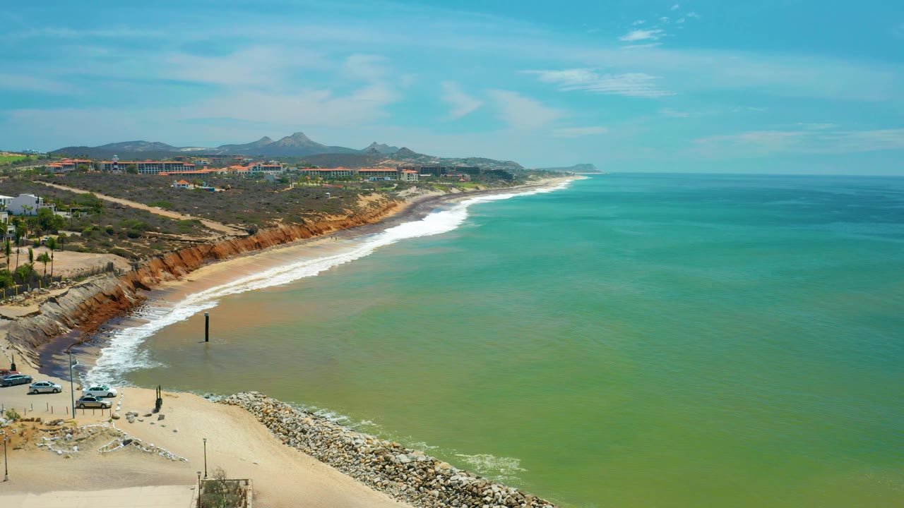 San Jose del Cabo shoreline from above with a broad field of green water, white surf tracing the beach, rust bluffs to the left, small distant buildings, mountains, and no visible people.