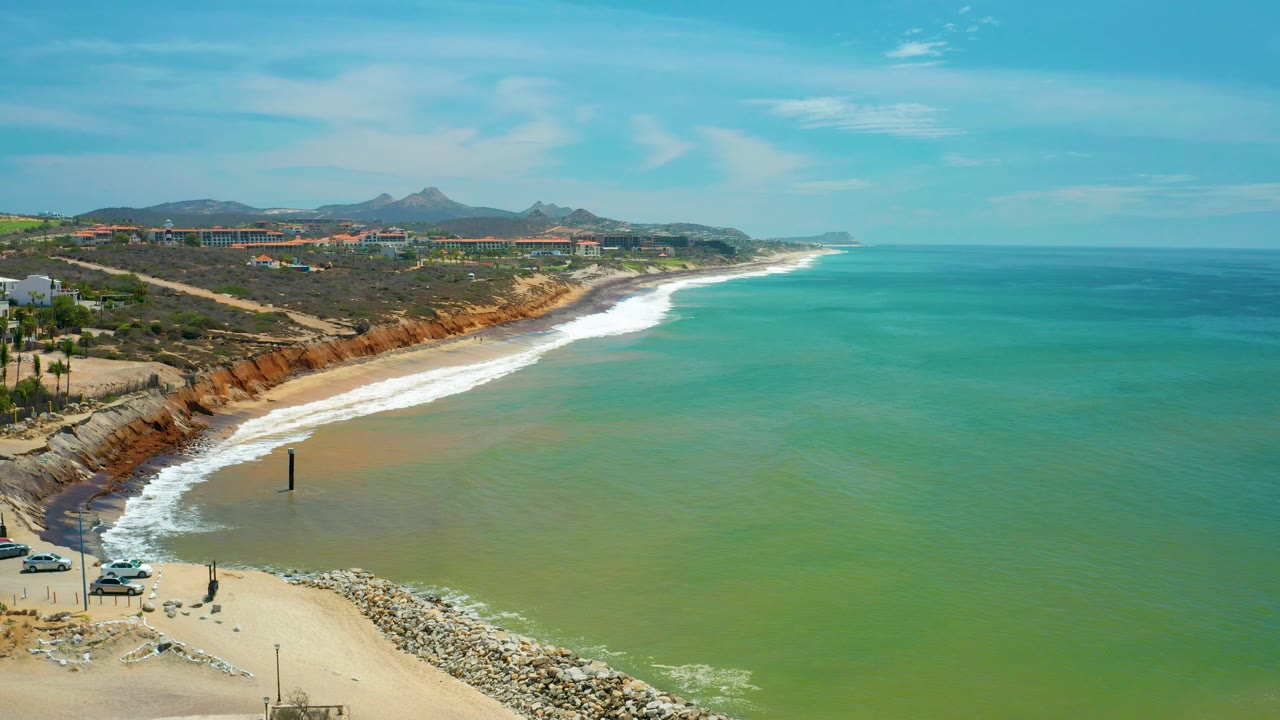 Elevated San Jose del Cabo view with waves breaking diagonally along the sand, green water filling the right side, bluff edges and buildings on the left, distant mountains, and no visible people.
