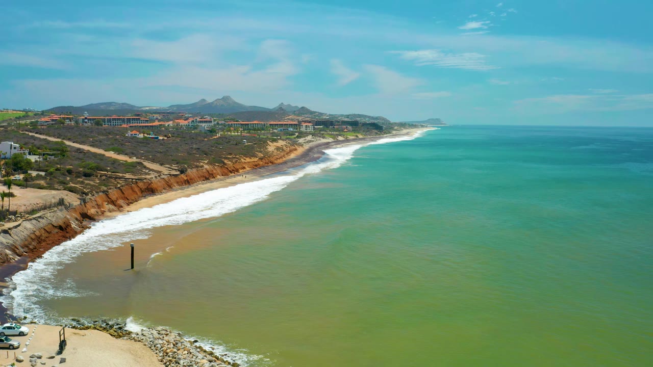 San Jose del Cabo coastal aerial with the surf line slightly farther along the beach, green-blue water spreading across the foreground, distant buildings and mountains, and no visible people.