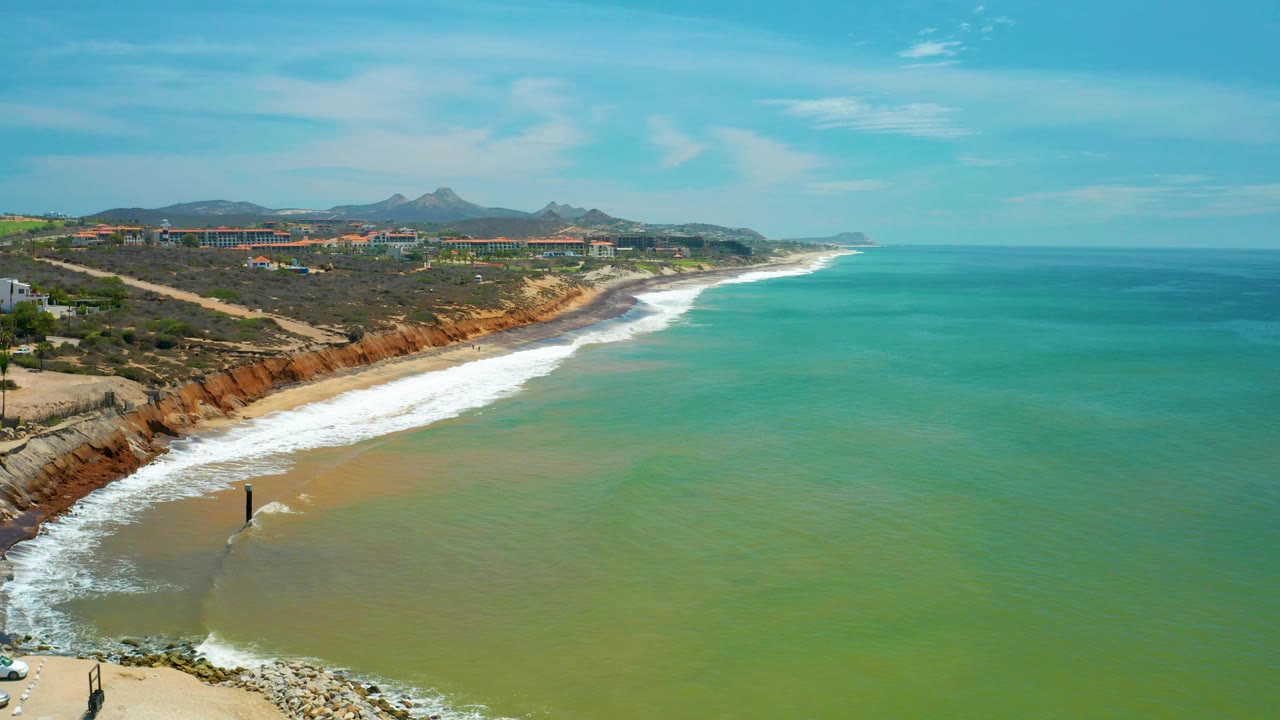 Wide San Jose del Cabo beach view with the dark vertical water structure nearer the lower left, white water fanning across the sand, offshore green water, background buildings, and no visible people.