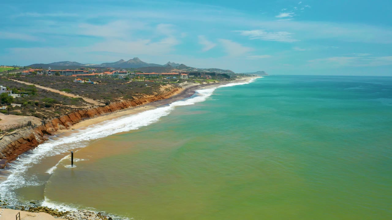 Aerial San Jose del Cabo coastline with waves breaking along the left beach edge, reddish cliff faces, open blue-green sea to the right, distant development, mountains, and no visible people.