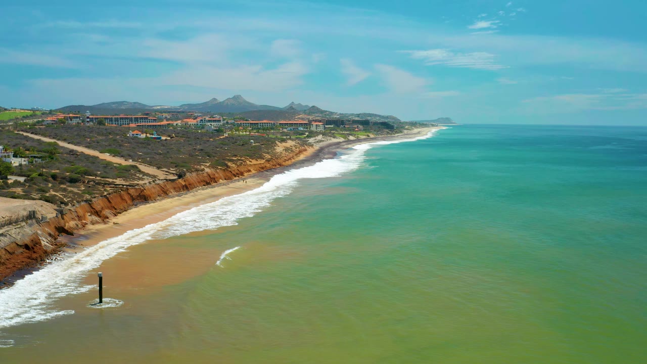 San Jose Del Cabo shoreline seen from above as white waves wash the sand below low planted bluffs, with green-blue water and no visible people.