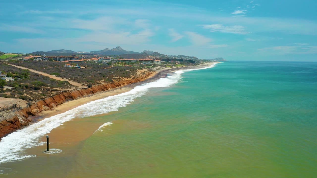 A diagonal San Jose Del Cabo beach line separates green-tinted sea water from reddish coastal bluffs and inland hills, with no visible people.
