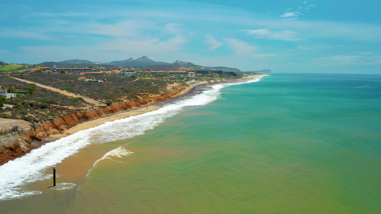 White surf curves into the sandy San Jose Del Cabo beach below brown bluffs, with turquoise water offshore, distant terrain, and no visible people.