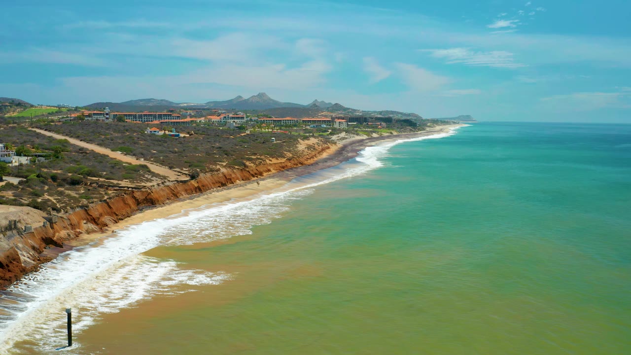 Elevated view over San Jose Del Cabo beach where white foam curves along the sand below rust cliffs, with turquoise water, distant buildings, low mountains, blue sky, and no visible people.