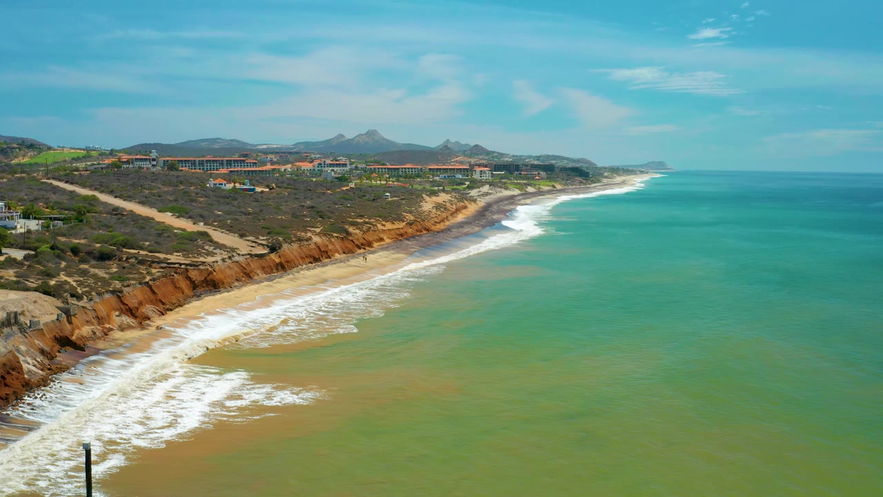Wide aerial frame of San Jose Del Cabo coastline with the cliff line and beach stretching into the distance, green ocean covering the right half, a level horizon, and no visible people.