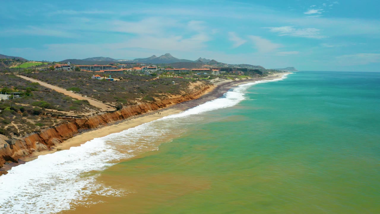 Aerial view in San Jose Del Cabo showing rust-colored cliffs, a sandy beach, white surf, green-blue water, inland roads and low buildings, with no visible people.
