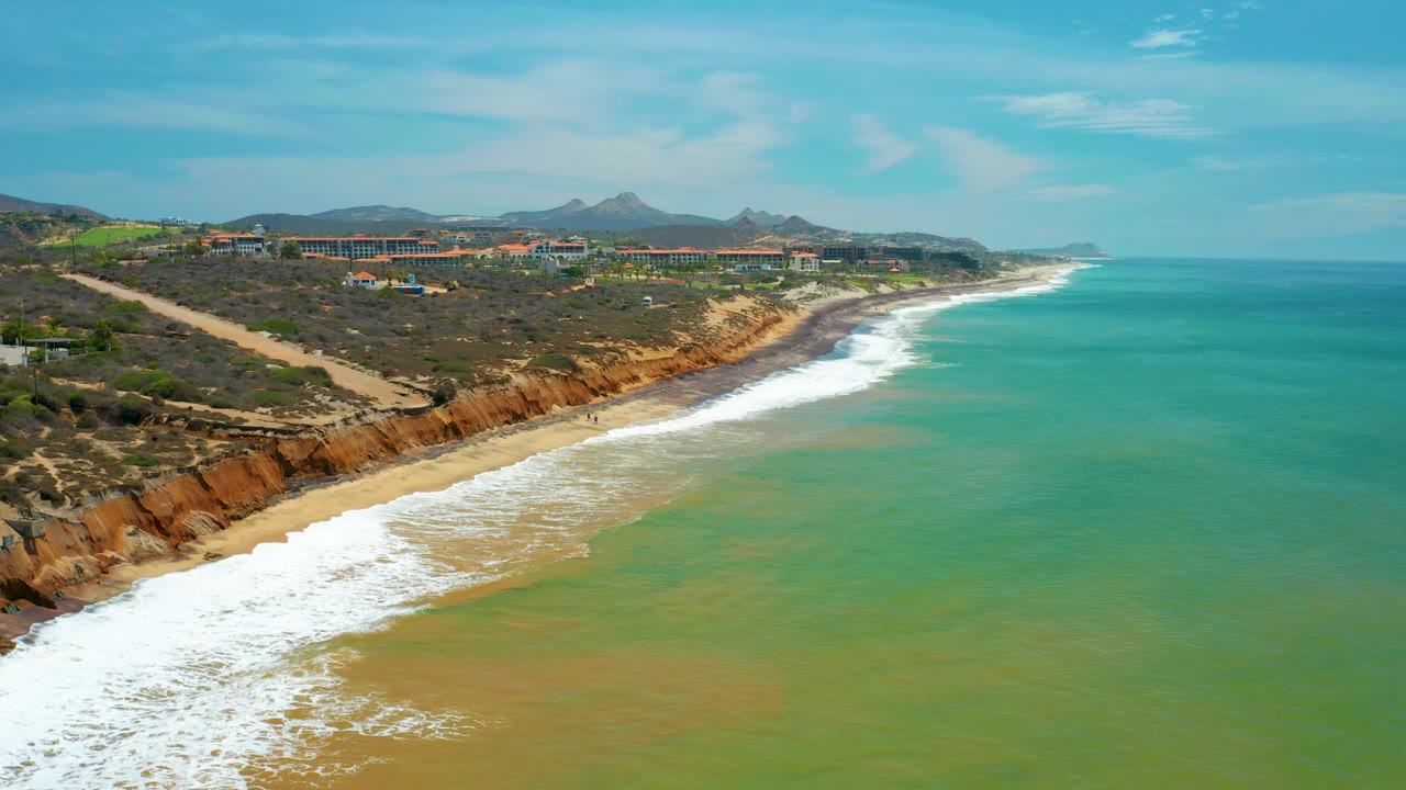 San Jose Del Cabo aerial view with open green-blue water filling the right side, a narrow sandy beach below orange-brown cliffs, inland roads and buildings above, and no visible people.
