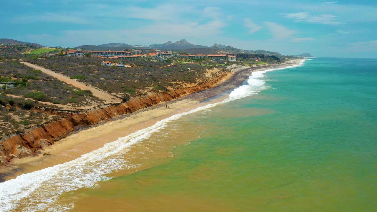 Aerial frame of San Jose Del Cabo shoreline with green-blue water in front, foamy surf tracing the beach, cliffs behind the sand, and no visible people.
