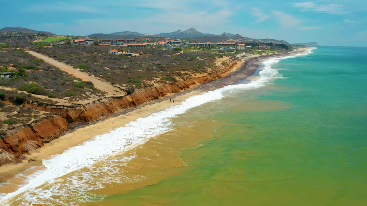 Wide aerial coastline with turquoise water, breaking waves, beach, and blufftop buildings.
