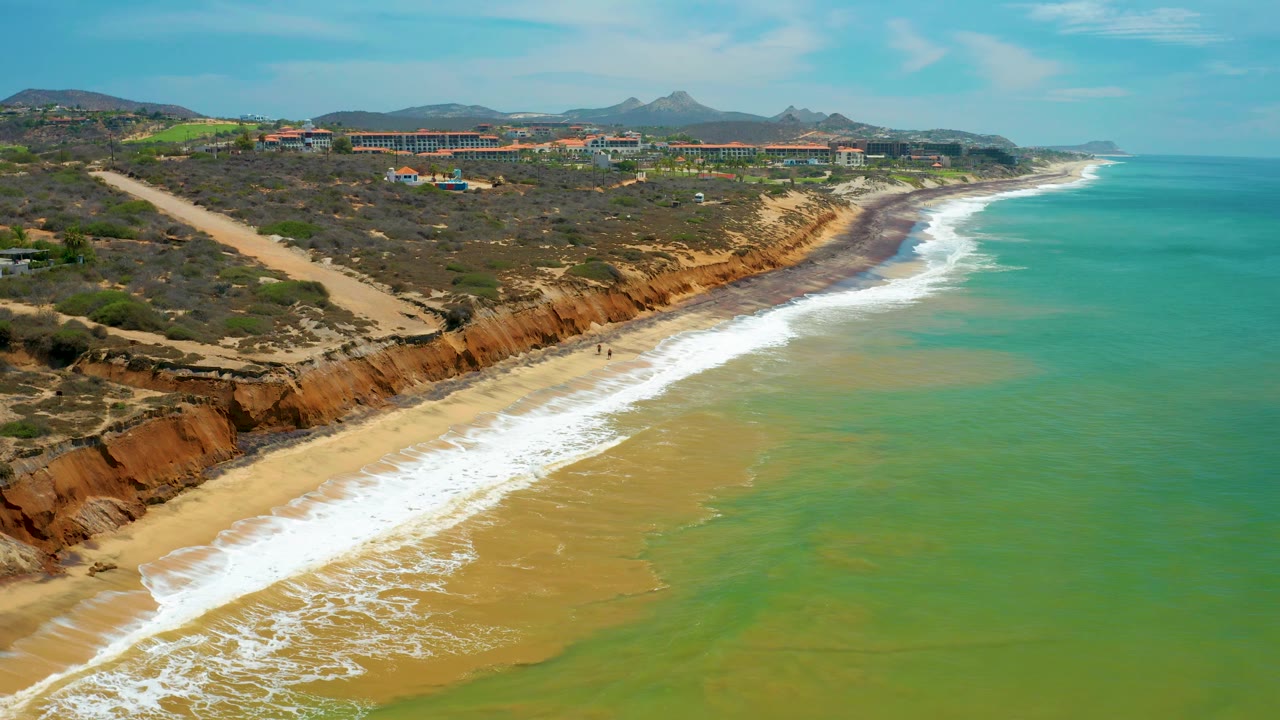 Aerial coastal frame in San Jose del Cabo with more open water to the right, a curved white surf band, pale sand below orange bluffs, inland structures, and no visible people.