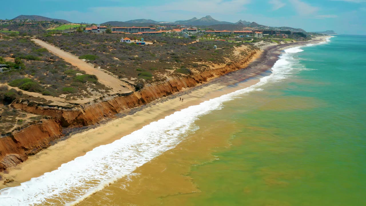 Aerial frame from San Jose del Cabo showing the breaking wave line farther along the beach, with the ocean on the right and dry hills above the cliffs, no visible people.