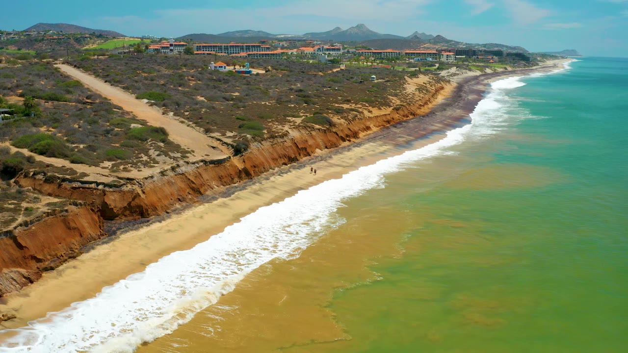 Aerial view over San Jose del Cabo with foam bands reaching the beach, a broad green-blue ocean surface, rust bluffs, and low inland development, no visible people.