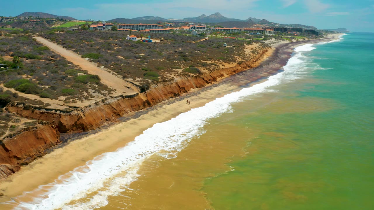 Mid-clip aerial frame in San Jose del Cabo where the surf curls across the lower beach and the cliff edge leads toward inland hills, no visible people.