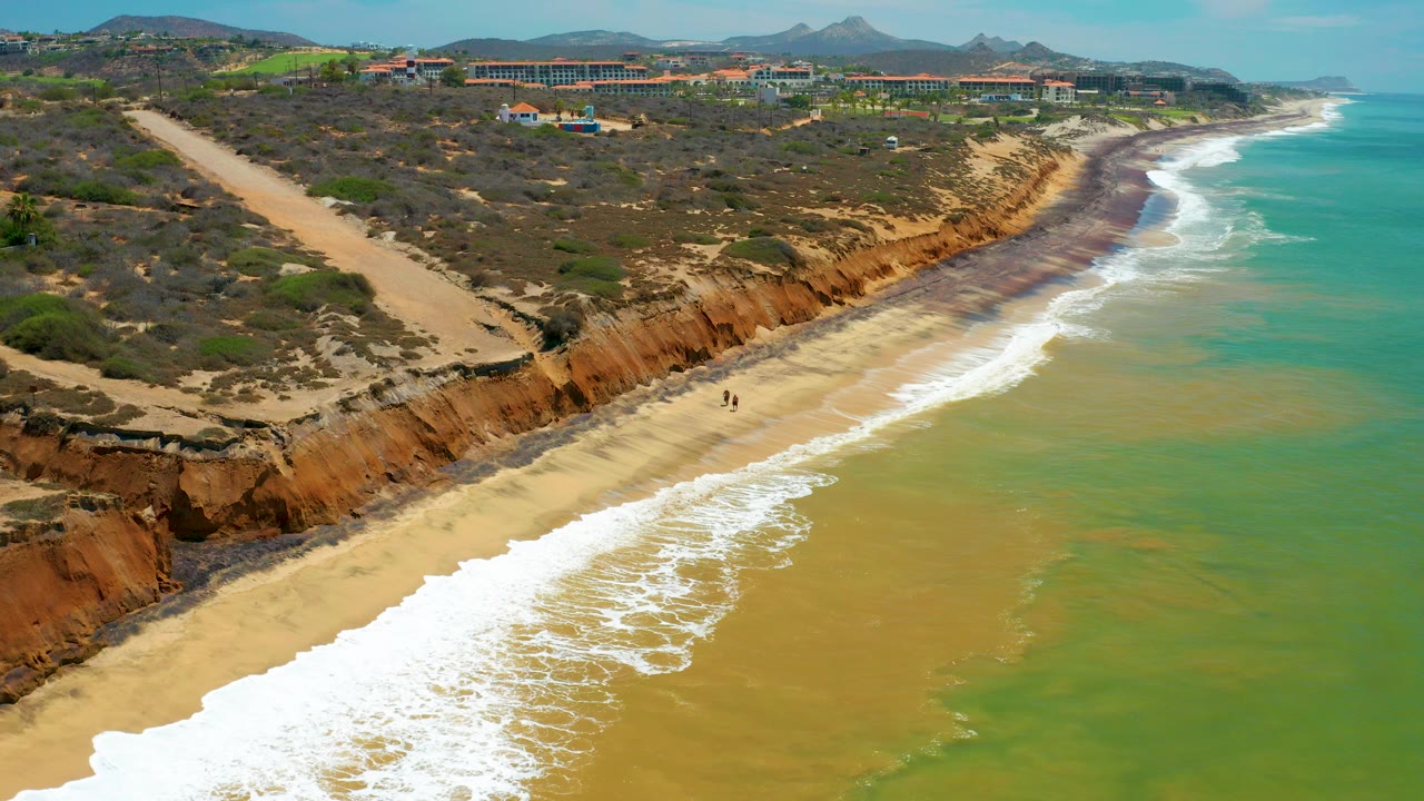 Aerial view over a San Jose Del Cabo beach with white surf reaching golden sand, rust-colored bluffs, scrubland, distant buildings, mountains, and tiny beachgoers near the shoreline.