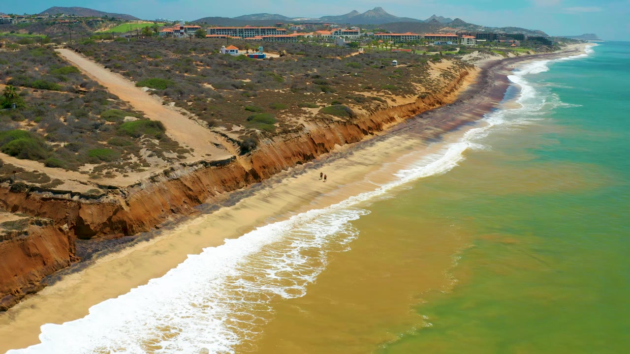San Jose Del Cabo coastline seen from above, with a broad band of pale surf across the lower beach, orange-brown cliff faces, scrub-covered land, distant resort-like buildings, and tiny people on the sand.