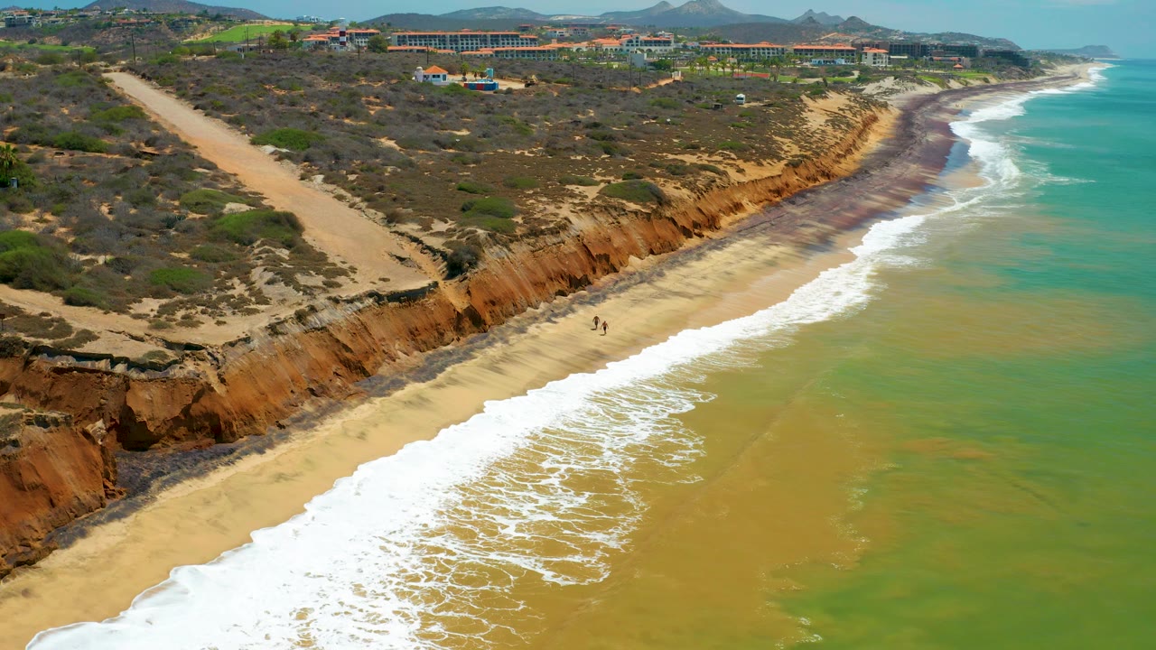 Aerial beach scene with waves rolling below scrubby cliffs and distant buildings.