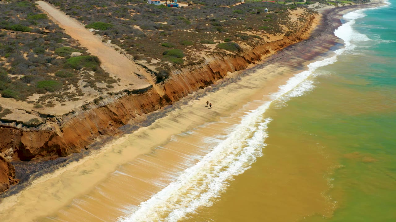 San Jose Del Cabo shoreline from above, with foamy waves advancing along tan sand below orange cliffs and two small walkers still visible near mid-beach.