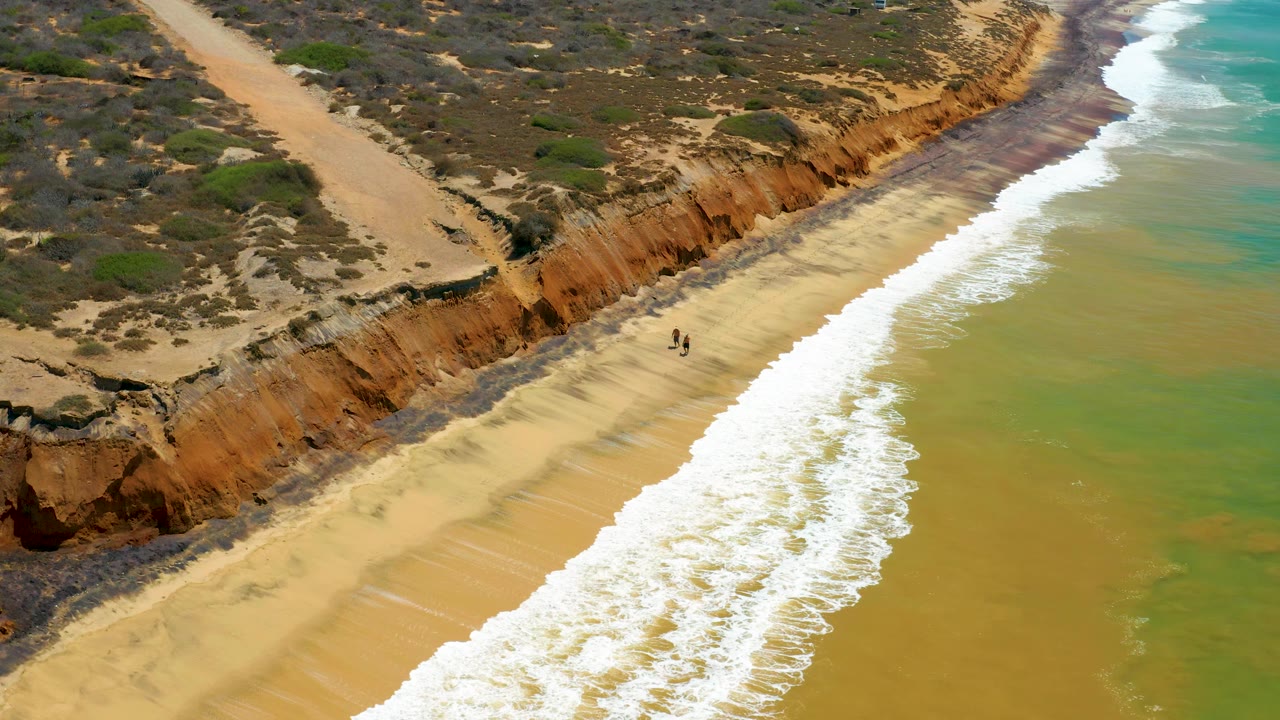 San Jose Del Cabo beach seen from an oblique drone angle, with a broad white surf band, tan sand, orange cliff face, scrub above it, and two distant walkers.