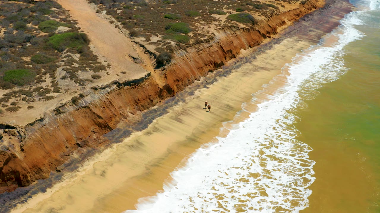 Aerial frame from San Jose del Cabo shows two small walkers on a pale beach below rust-colored cliffs, with white surf entering from the right.