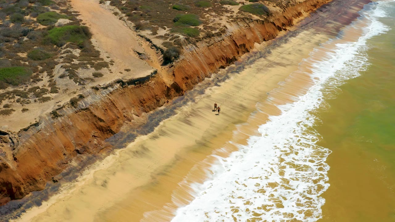 The two people appear higher on the beach in San Jose del Cabo, with a bright band of foam beside them and scrub-covered ground above the cliff edge.