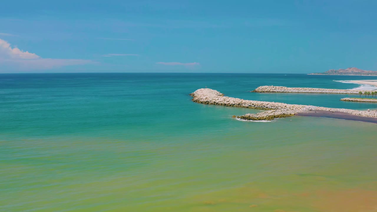 Elevated coastal frame from San Jose Del Cabo, Mexico, with turquoise water in front, pale rock jetties to the right, a small distant island, and no visible people.