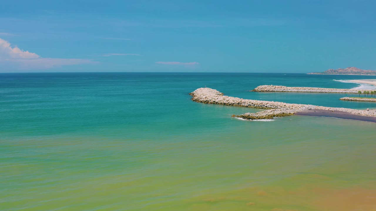 San Jose Del Cabo ocean view with blue water beyond green shallows, two pale rock arms extending from the right edge, distant land at far right, and no visible people.