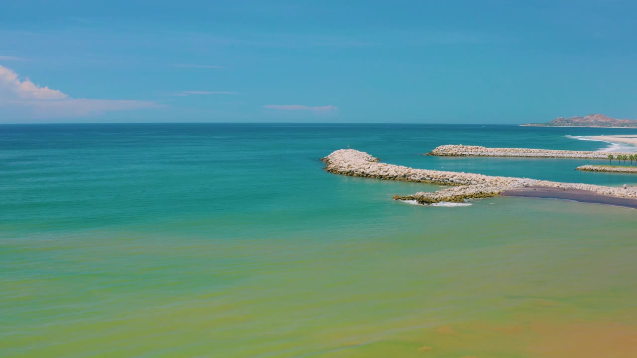 Wide San Jose Del Cabo coastal water frame with green surface tones in the foreground, a pale rock breakwater crossing the right side, blue sea beyond, and no visible people.