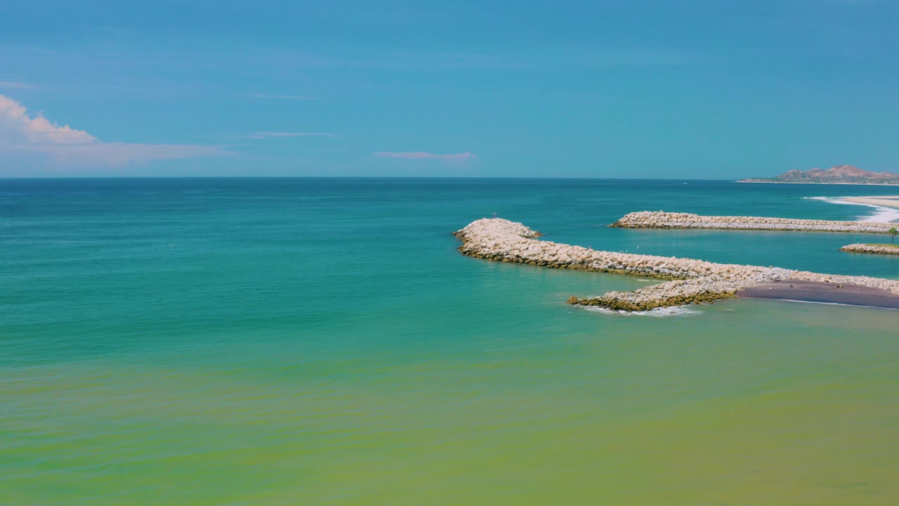 Opening frame of San Jose Del Cabo ocean with turquoise water, pale rock breakwaters crossing the midground, distant shoreline on the right, and no visible people.