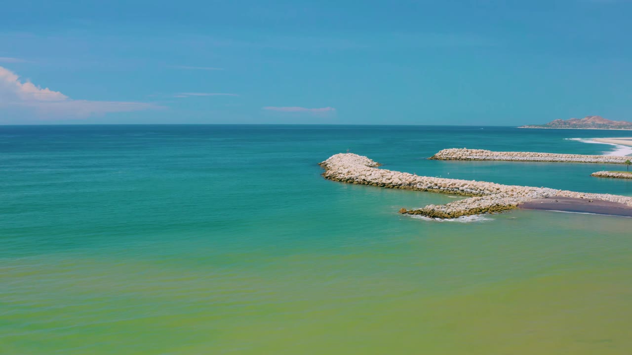 San Jose Del Cabo shoreline view with a long pale rock path extending from the lower right toward the center, blue-green ocean around it, and no visible people.