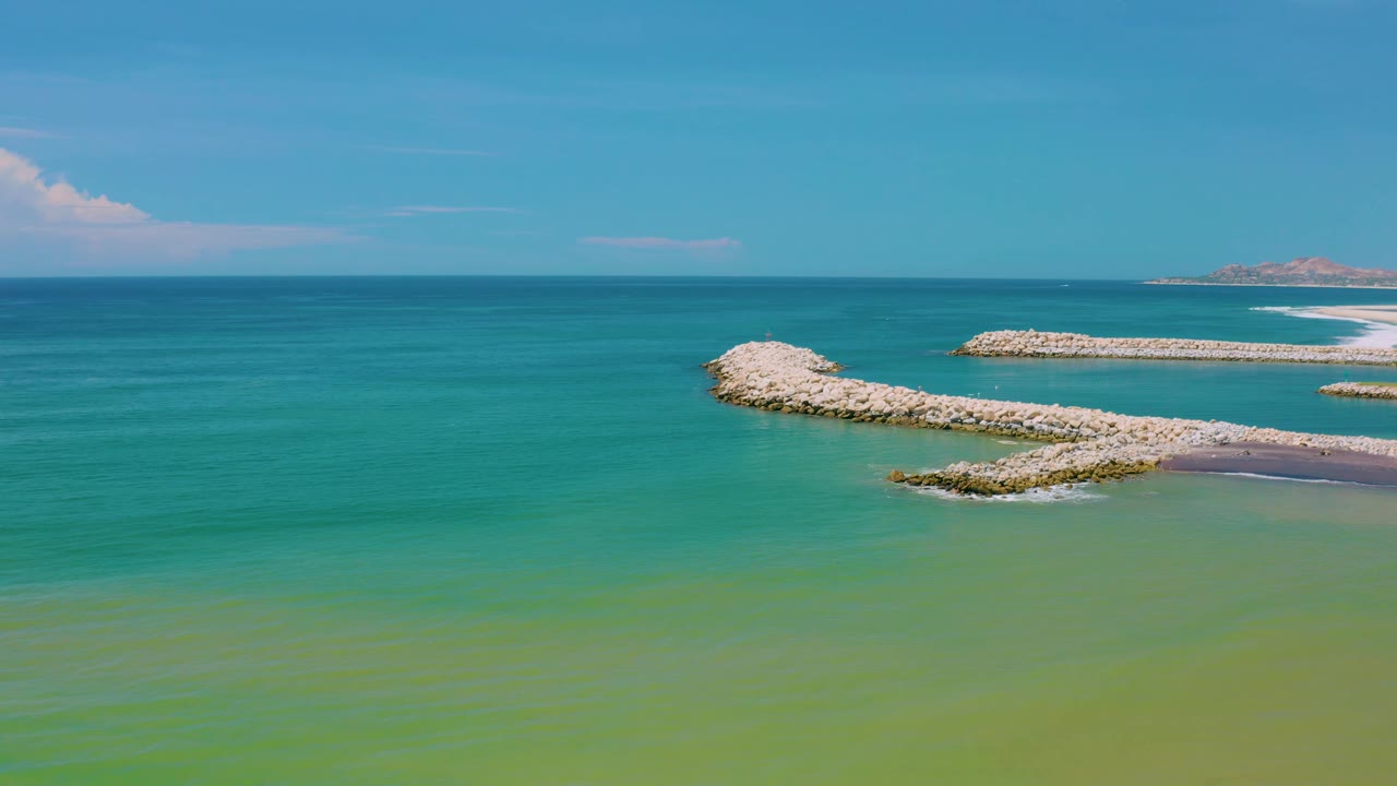 Aerial-looking San Jose Del Cabo frame with curved breakwater rocks near center right, calm turquoise water across most of the image, and no visible people.